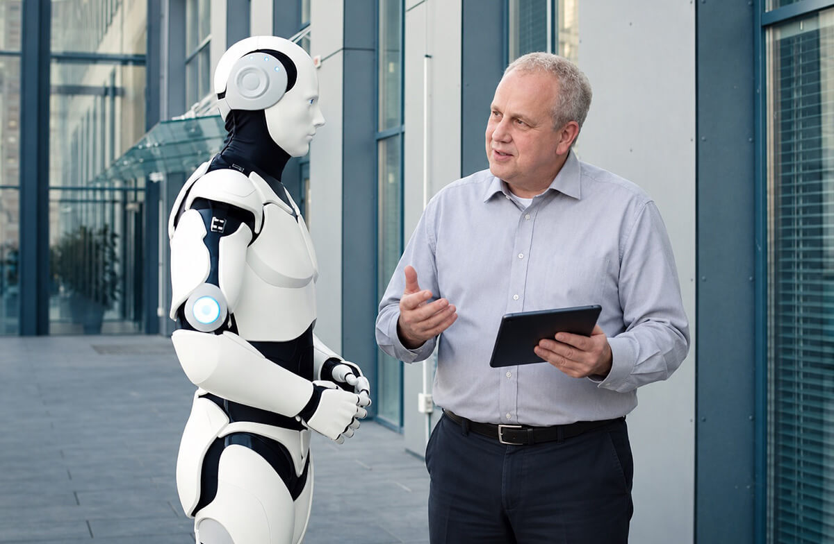 RoboterFlüsterer Kent Schwirz mit Tablet neben einem humanoiden Roboter vor einem Betriebsgebäude
